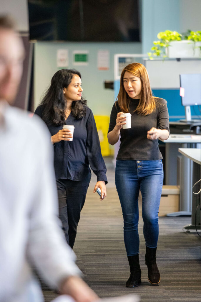 Two women walk through an office whilst holding paper cups, engaged in conversation about designing across time zones. Desks, office equipment, and a computer monitor are visible in the background, highlighting the collaborative environment typical of async teams at Learnosity.